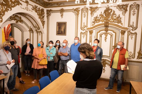 Eine junge Frau teilt den anwesenden Mitmachern einige interessante Aspekte zur Schlossgeschichte mit. Die Besucher bestaunen den weiß-golden verzierten Schlossraum und die Decke.