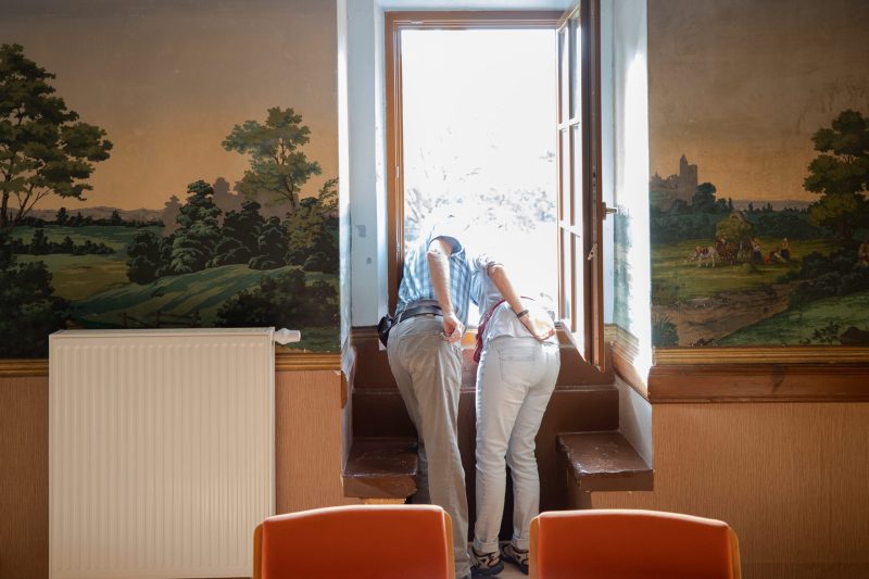 Zwei Schlossbesucher lehnen sich sich weit zu einem Fenster hinaus. Rechts und links von ihnen sind Ausschnitte der kostbaren Tapete mit Landschaftsbildern zu sehen.