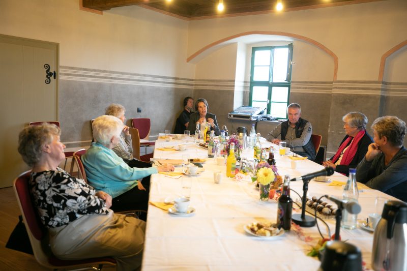 Blick auf eine lange Tafel in einem niedrigen, historisch anmutenden Raum. Daran sitzen links und rechts jeweils drei Personen, an der Stirn sitzt die Moderatorin. Die Tafel ist mit Kuchengeschirr gedeckt. Dazwischen stehen Mikrofone.