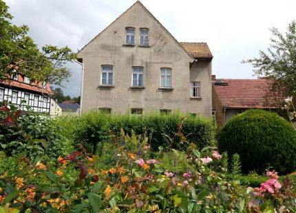 Blick aus dem bunten Blumengarten auf das Geburtshaus von Alfred Ahner in Wintersdorf. Das dreigeschossige Haus hat ein gelbes Ziegeldach. Die grau-braune Fassade und die weißen Sprossenfenster sind unsaniert und lassen Witterungseinflüsse erkennen.